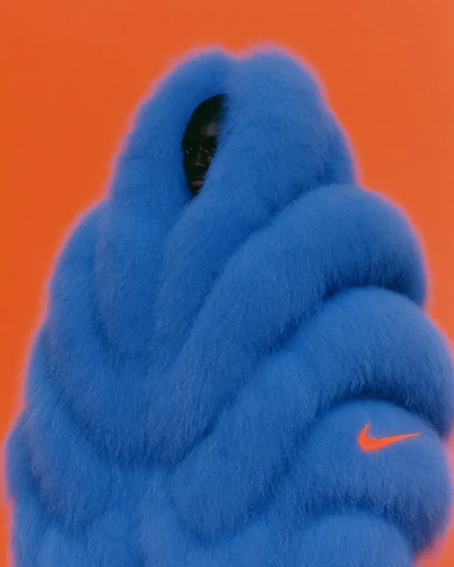 A full-figure portrait of a person enveloped in a vibrant blue, fluffy garment, set against a bold orange background. The photograph captures a surreal and playful mood, highlighting the contrast between the garment and backdrop.