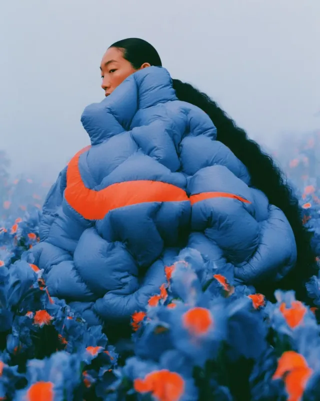 A full-figure portrait of a woman enveloped in a vibrant, oversized blue puffer coat with orange accents, set amidst a field of striking blue flowers. The image employs a surreal and dreamy mood with a soft and hazy atmosphere.
