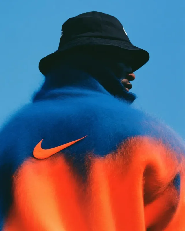 A striking editorial photograph featuring a man in a colorful, oversized, gradient sweater that blends orange and blue hues, complemented by a black bucket hat. The mood is vibrant and bold, with a clear blue sky as the backdrop.