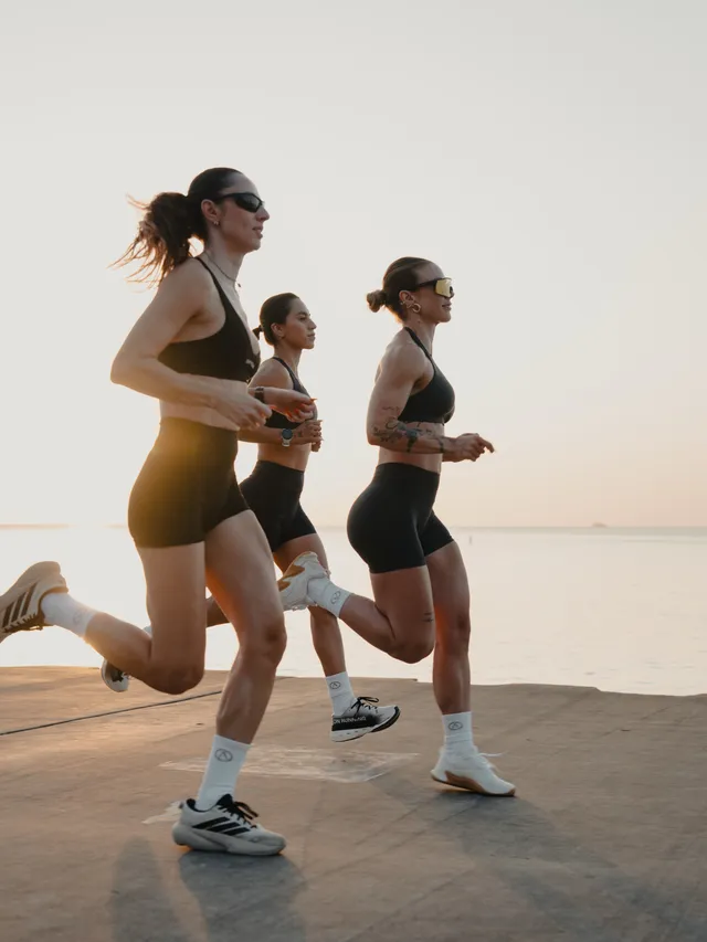 A dynamic editorial photograph featuring three women running along a waterfront at sunset. The soft golden hues of the sunset contrast with their dark athletic gear, creating an energetic and inspiring mood.