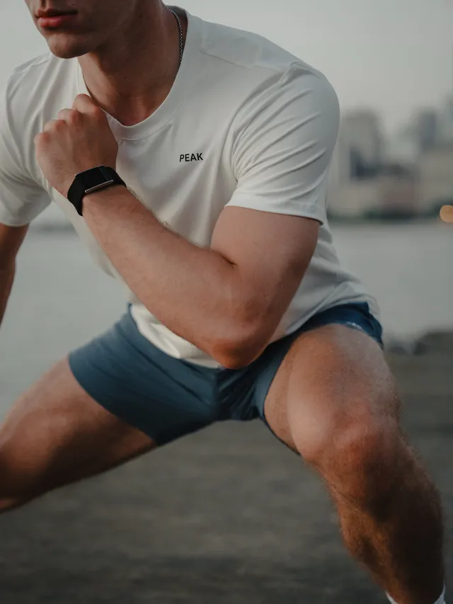 A man in a white athletic shirt and blue shorts performs a squat, captured in an editorial photography style. The scene is set during sunset, creating a warm and dynamic mood with a palette of soft blues and soft oranges.