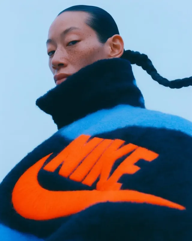 A dramatic editorial photograph of a woman in a high-neck sweater with a prominent logo, conveying an intense and confident mood. The dominant colors are blue, black, and bright orange.