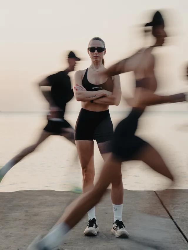 A dynamic editorial photograph featuring a woman standing confidently in the foreground while blurred athletes in motion pass by her. The mood communicates determination and energy, with a color palette dominated by soft pastels against a calm water backdrop.