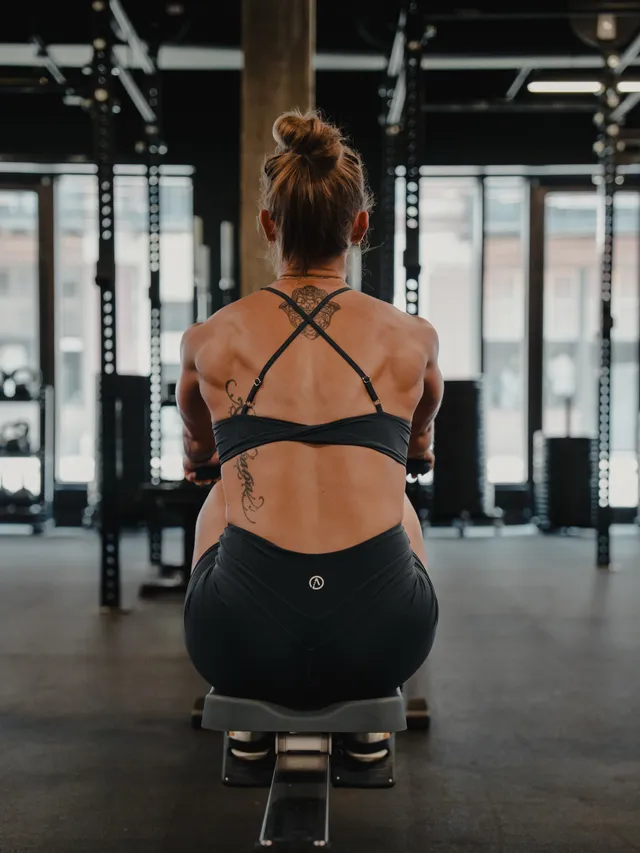A focused portrait of a woman from the back, captured in a gym setting. The medium is editorial photography, showcasing a moody atmosphere with a dark color palette dominated by blacks and grays.