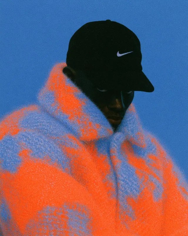A moody editorial photograph of a man wearing a colorful oversized sweater and a cap. The image features a striking blue background with bold orange patterns, creating a vibrant contrast.