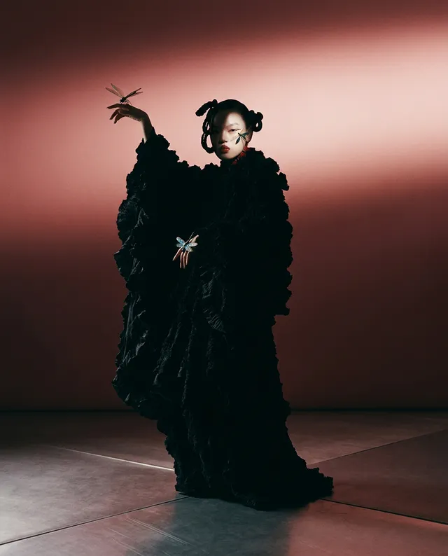 A full-figure portrait of a woman in a dramatic, ruffled black gown, captured in a high-gloss editorial photograph. The scene conveys a moody atmosphere with soft, directional lighting highlighting the textures of the fabric against a warm, reddish background.