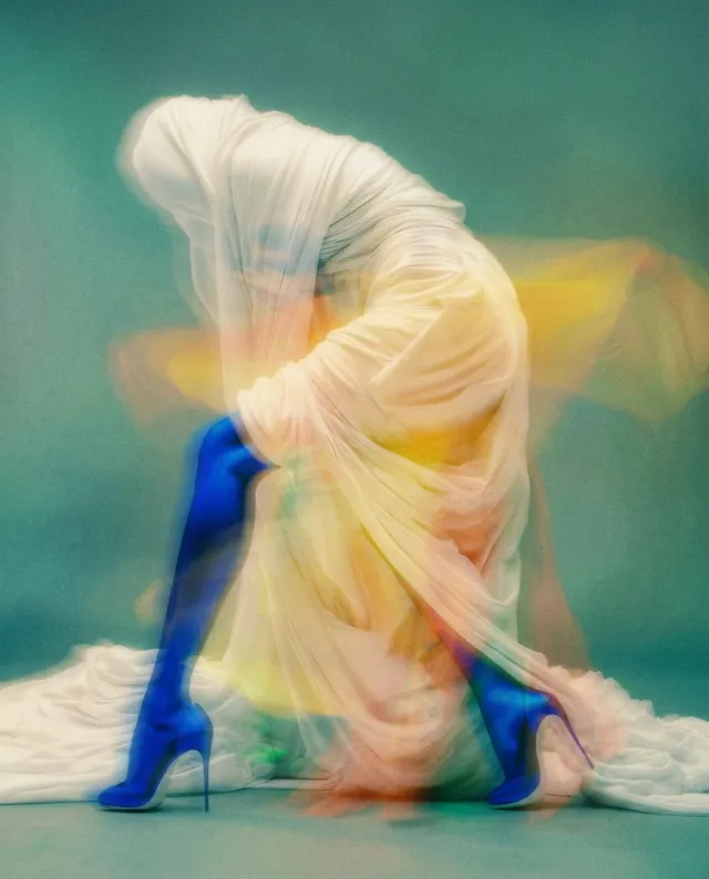 A distorted figure draped in flowing, iridescent fabric, conveying movement and abstraction. The image showcases vibrant colours like blue, yellow, and orange, with a dreamy, surreal quality.