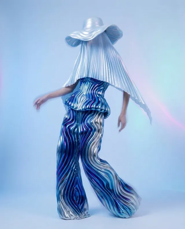 A full-figure portrait of a woman wearing a sculptural, iridescent outfit that showcases flowing lines and texture. The lighting is ethereal with a cool color palette of blues and silvers, creating a dreamy and futuristic mood.