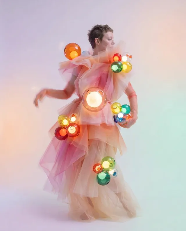 A whimsical portrait of a woman draped in flowing fabric adorned with colorful, translucent spheres. The visual medium is digital illustration, evoking a playful and surreal mood with soft pastel hues.