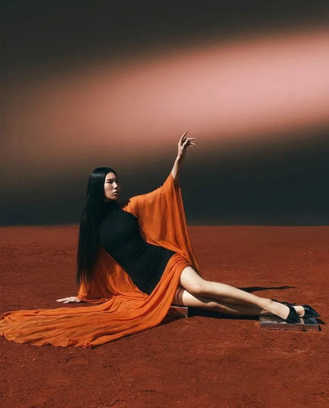 A woman elegantly posed in a flowing orange dress that contrasts with a rich reddish-brown background, capturing a dramatic editorial style. The mood is striking, emphasizing bold colors with soft lighting.