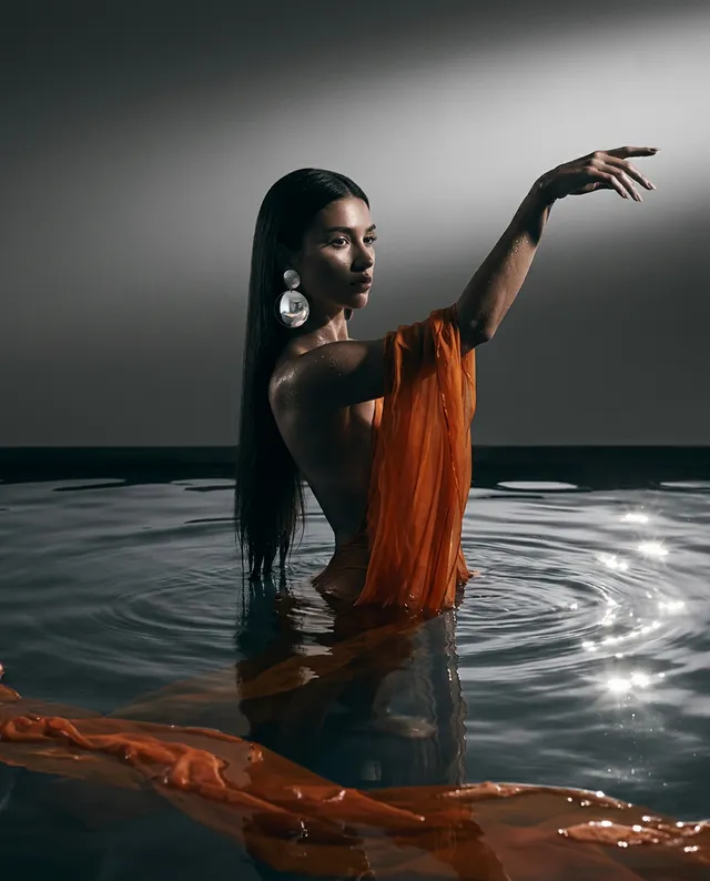 A striking portrait of a woman partially submerged in water, draped in flowing orange fabric. This high-contrast editorial photograph features dramatic lighting and a serene but moody atmosphere, with silver accents reflecting in the water.