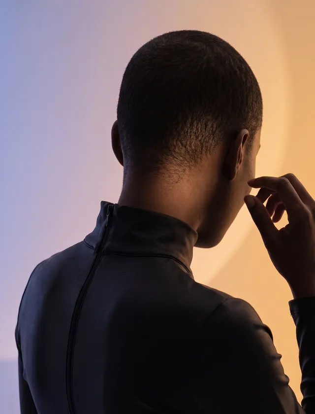 A close-up portrait of a man seen from the back, wearing a sleek, high-collared garment. The stylish editorial photograph features a gradient background transitioning from soft orange to muted purple, enhancing the contemplative mood.