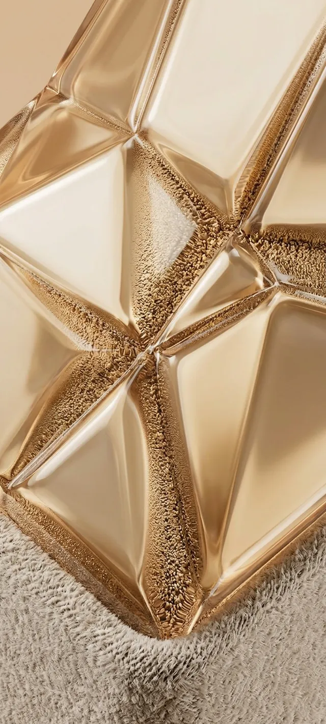 A close-up of a sculptural gold object with geometric facets reflecting light, set on a soft textured surface. The composition showcases a blended color palette of gold tones and soft neutrals, exuding a luxurious mood.
