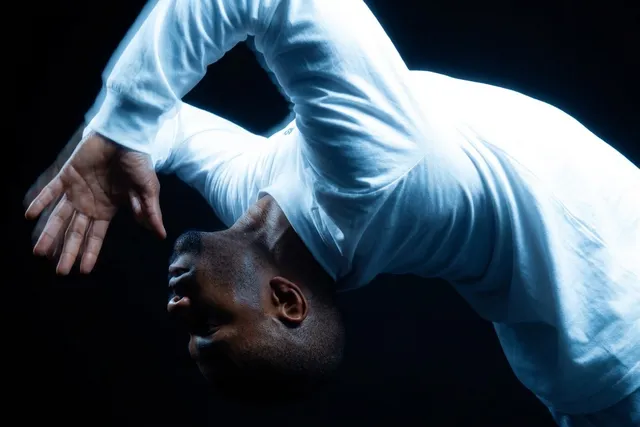 A full-figure portrait of a man performing a dance move, captured in dramatic lighting, emphasizing movement and fluidity. The image is characterized by a moody atmosphere and a dominating blue hue against a dark background.