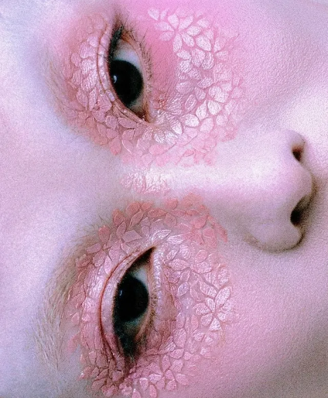 A close-up portrait of a woman's face featuring delicate floral patterns across her eyelids, captured in a dreamy, ethereal style. The overall mood is surreal, with soft pink hues dominating the image.