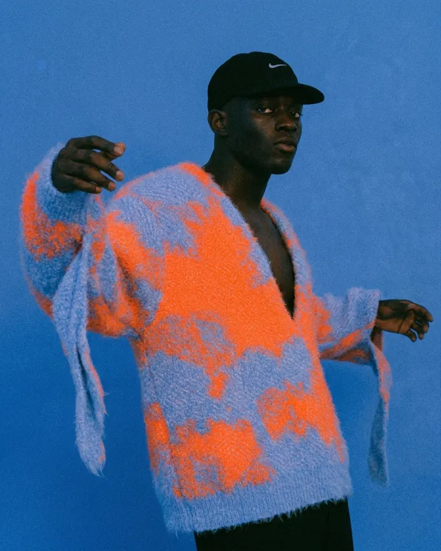 A man wearing a vibrant orange and blue fuzzy sweater against a blue background.