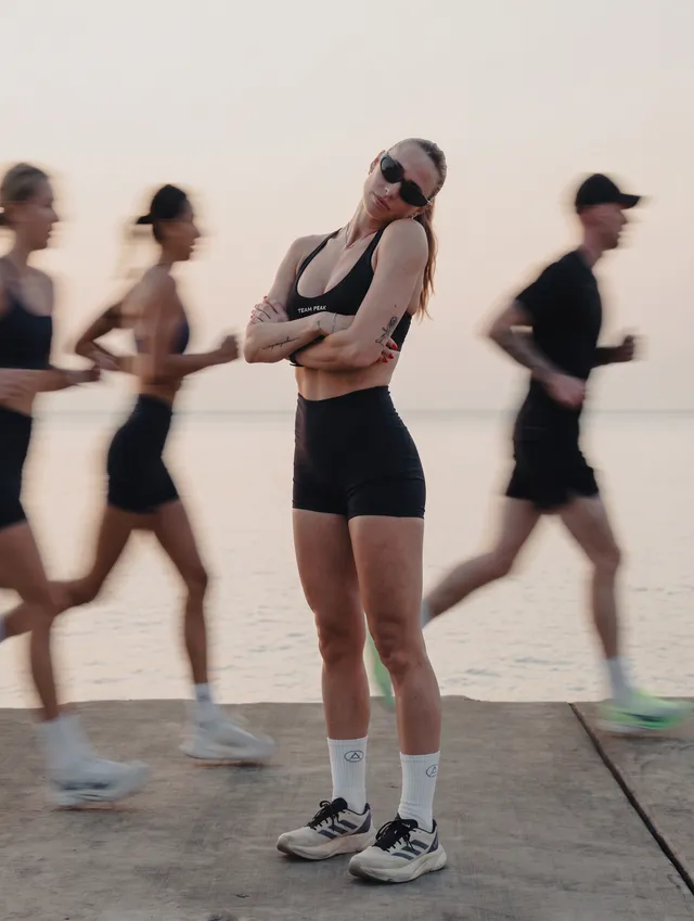 A confident woman stands in front of a calm water body with blurred runners in the background.