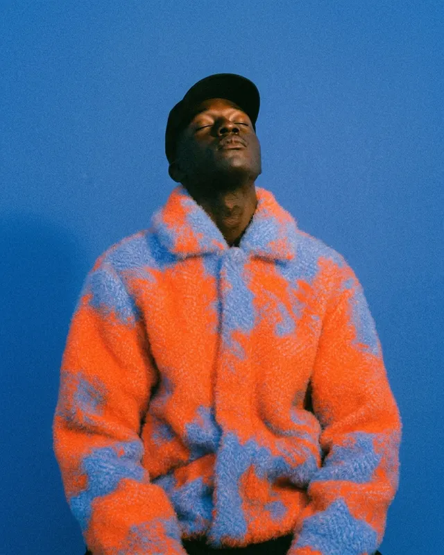A man wearing a vibrant oversized fuzzy jacket stands against a bright blue background.