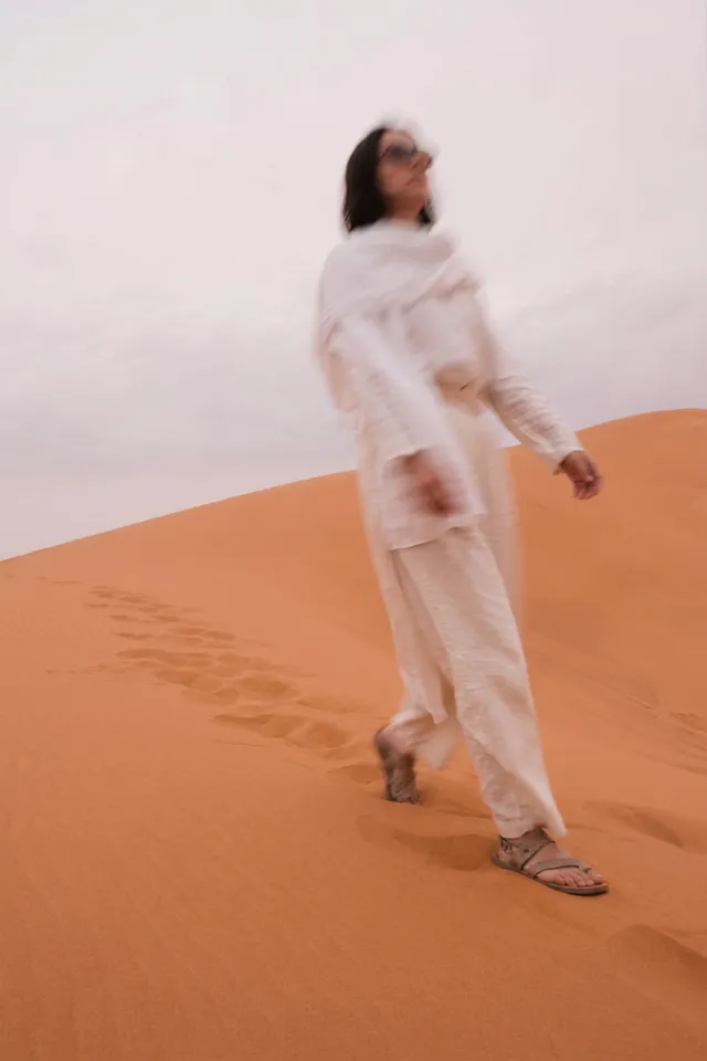 A woman walking through a sandy desert landscape, captured in a soft, editorial photography style. The mood is serene, with neutral and warm tones dominating the scene.