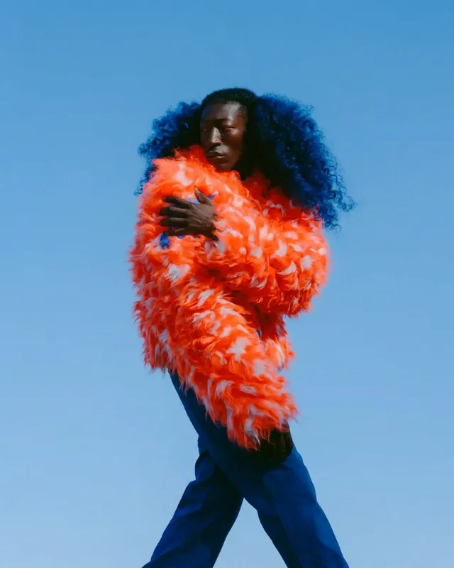 Full-figure portrait of a model wearing an orange feathered coat with a blue sky background.