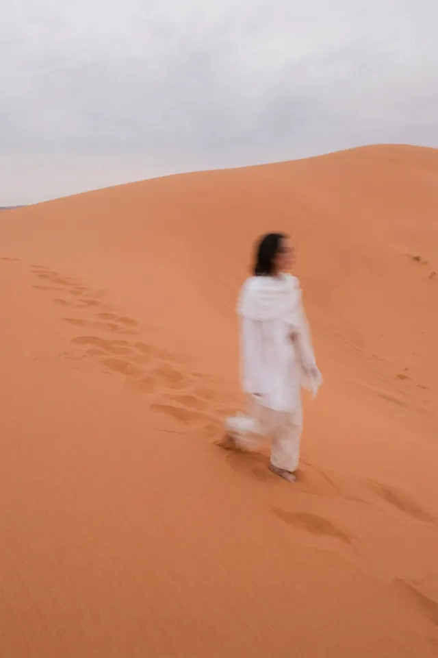A blurred figure of a woman walking across a vast orange sand dune, captured as an editorial photograph. The scene conveys a serene and moody atmosphere, with soft, muted tones dominating the image.