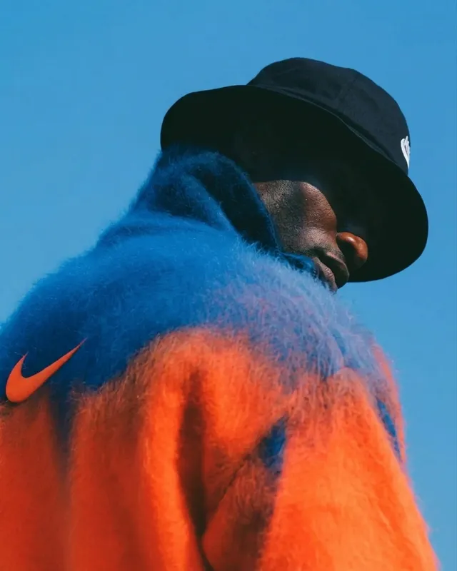 A man in an orange and blue fur jacket and black bucket hat with a blue sky background.