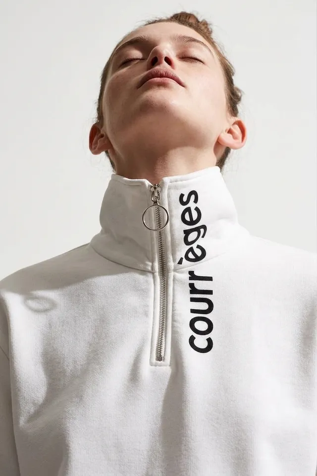 A portrait of a woman wearing a zippered white sweatshirt with black branding. The image captures a fashion editorial style with a clean, minimal background, emphasizing the garment and the model's expression.