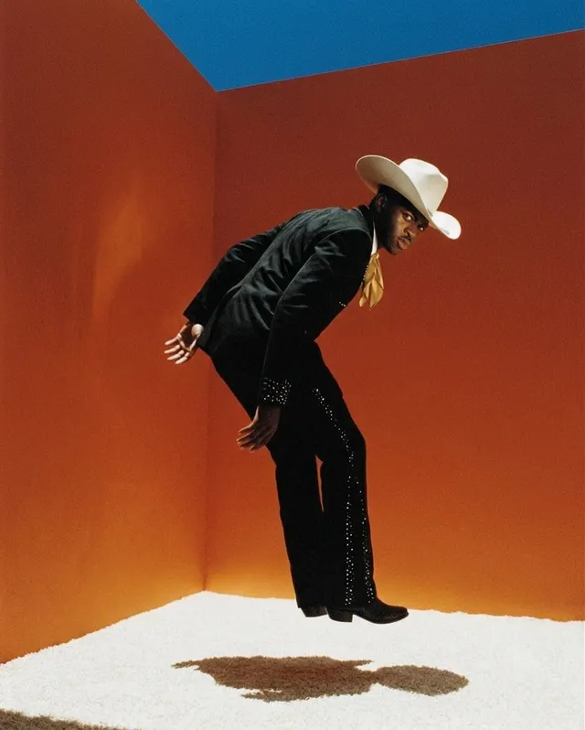 A man dressed in a black suit and cowboy hat, mid-jump against an orange backdrop, conveying a playful and energetic mood. The scene is captured in a high-gloss editorial photograph style.