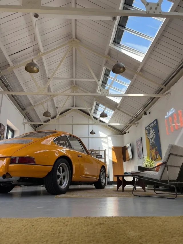 An editorial photograph capturing a vintage orange sports car inside a spacious, modern loft. The mood is bright and inviting, with natural light flooding through skylights, complemented by neutral colors of the interior and the striking orange of the car.