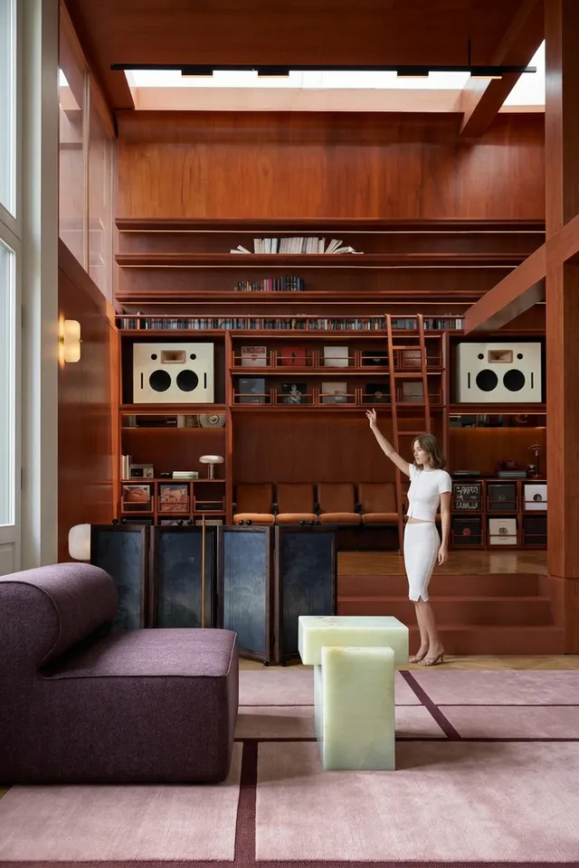 A woman in a fitted white dress interacts with a modern interior, featuring rich wooden walls and sleek furniture. The style is editorial photography, capturing a blend of sophistication and minimalism, with a focus on warm wood tones and soft pastels.