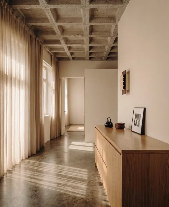 A serene interior space featuring minimalist design, with light filtering through sheer curtains. The dominant colors are warm neutrals and soft browns, evoking a calm ambiance.