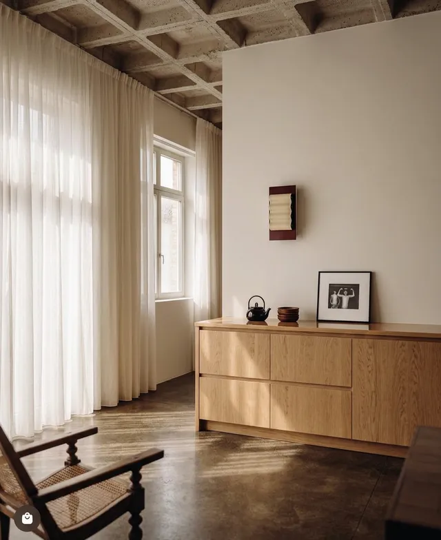 A cozy interior scene featuring a wooden cabinet, framed artwork, and a kettle. The atmosphere is warm and inviting, highlighted by soft natural light filtering through sheer curtains, with a color palette of neutral tones.