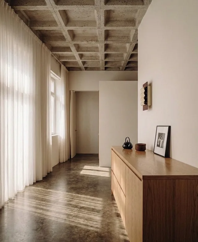 A serene interior space featuring minimalist design elements, characterized by soft natural light filtering through sheer curtains. The dominant colors are warm neutrals with a touch of wood.