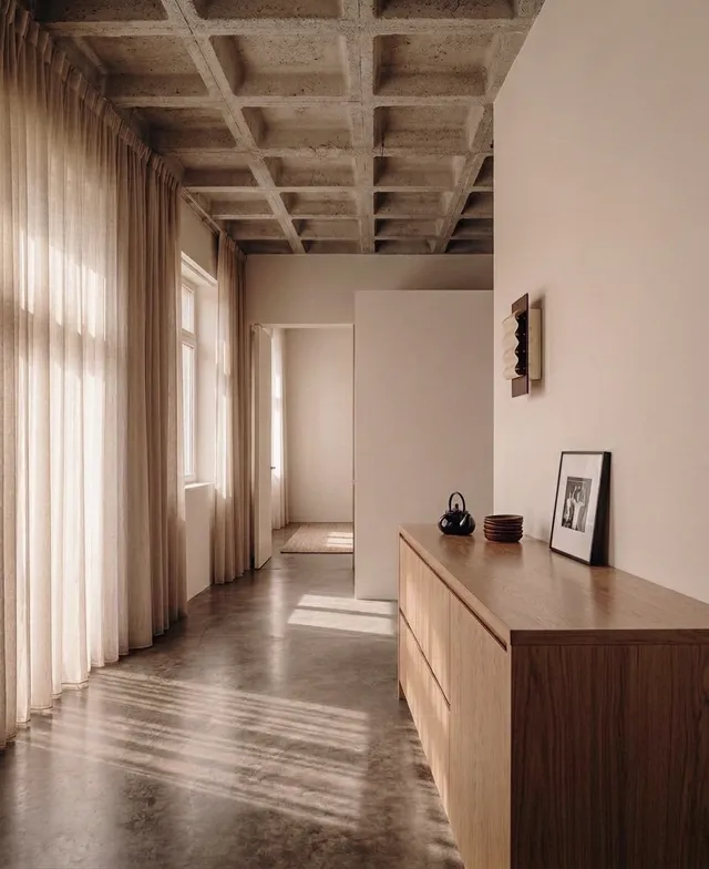 A serene and minimal interior space featuring smooth concrete floors and a wooden sideboard. The space is bathed in soft natural light, with flowing curtains adding warmth to the neutral color palette.