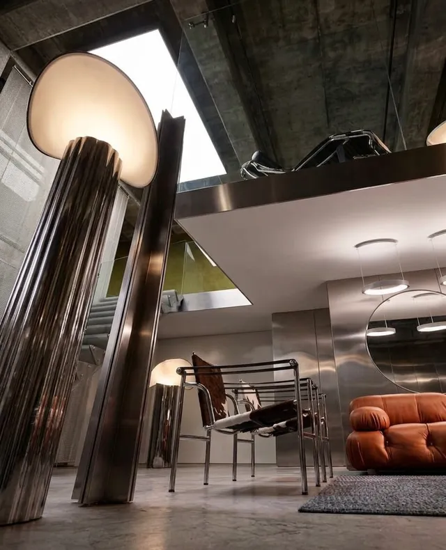 A modern interior space featuring sleek metallic furniture and minimalist design, captured as a high-gloss editorial photograph. The predominant colors are silver, gray, and warm orange tones from the sofa.
