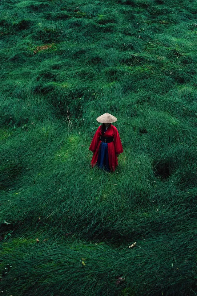 A woman standing amidst lush green grass, wearing a vibrant red robe and a traditional straw hat, creating a striking contrast with the surroundings. The scene captures an editorial photography style with a serene mood.