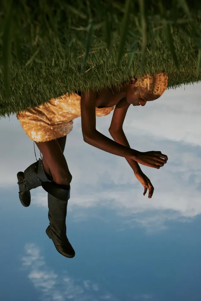 A woman in a patterned yellow dress, captured in an unconventional upside-down pose, emphasizes a surreal atmosphere. The editorial photography features earthy tones of green grass and a blue sky, creating a striking contrast.
