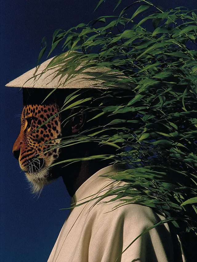 A portrait of a man with a leopard's face, surrounded by green foliage. The image is a vibrant editorial photograph, characterized by striking contrasts and deep colors.